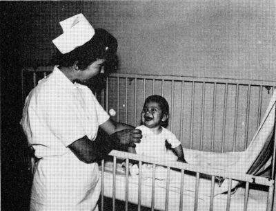 An infant sits in a crib and smiles at a nurse who is propping him upright