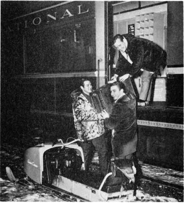 Two men lift a box onto a train. A man stands on the train and reaches for the box.