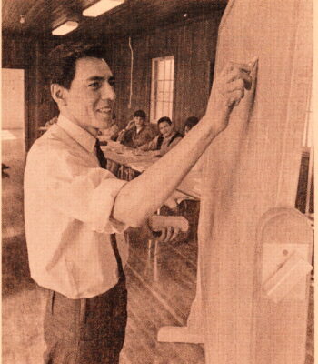 A man draws on a chalkboard. Other young men can be seen sitting in a row at desks behind him.
