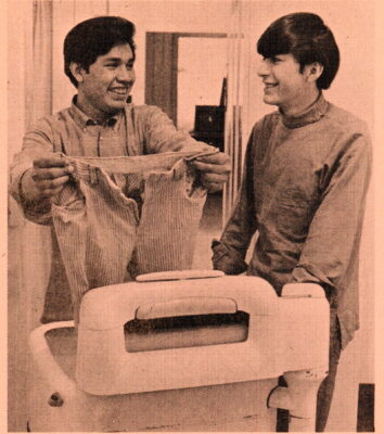 A young man smiles and holds up a pair of pants. Another young man looks at him and smiles.