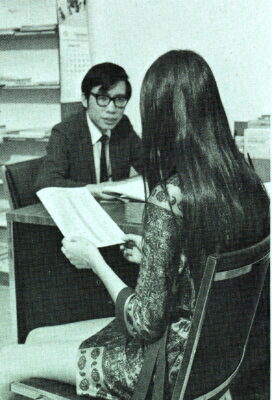 A woman sits in a chair facing away from view, reading a magazine. A man in a suit sits at a desk across from her.