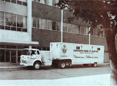 A truck with the words "Sanatorium Board of Manitoba Mobile Detection Unit" on the side sits next to a building.