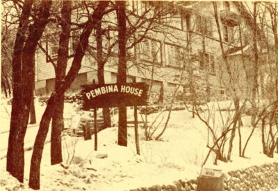 A sign that reads, "PEMBINA HOUSE" stands on the snowy ground amongst trees. A building stands in the background.