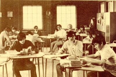 Ten young adults sit at desks scattered throughout a room with two large windows on the back wall.