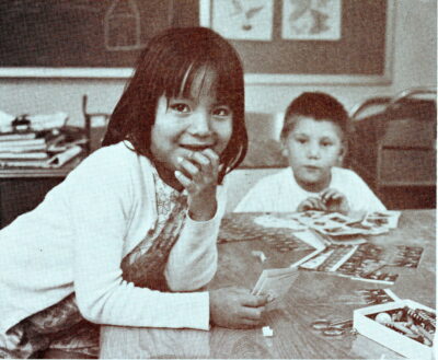 A boy and girl sit at a table in a classroom and play with stamps. The girl sits in the foreground with her elbows resting on the table and one hand at her mouth.