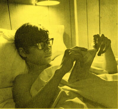 A young man lies on his back in bed with his head propped on a pillow, and reads a book. A light shines above him.