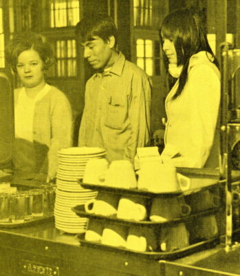 Three young adults line up in a cafeteria next to stacks of dishes.