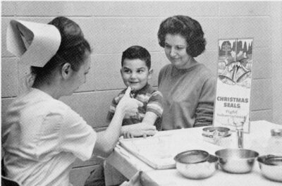 A nurse injects a child who sits on a woman's lap. A Chirstmas Seals sign sits on the table next to them.