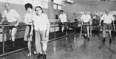 Five people with artificial legs practice walking. A nurse supports a woman.