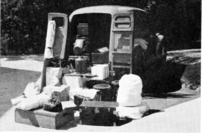 The back of a vehicle is opened, revealing boxes and equipment, spilling onto the ground in front of it.