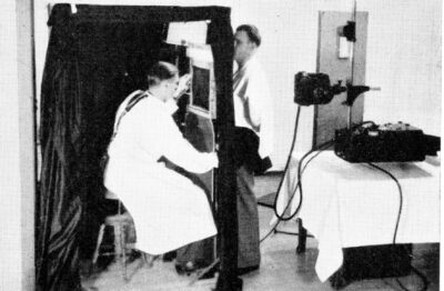 A man presses his chest against an x-ray plate as another man sits infront of him and operates the machine.