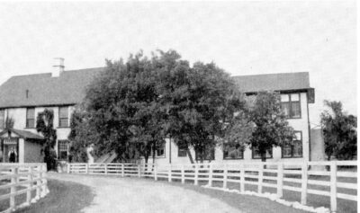 A curved road lined with a fence leads to two buildings. Trees stand in front of the buildings.
