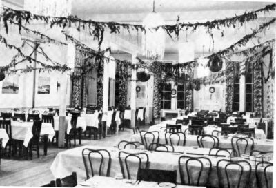 A room filled with multiple dining tables which are set with table cloths and dishes. Garland is strung from the ceiling.