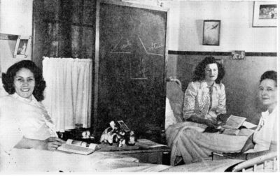 Two women sit in beds with books on their laps. Another woman sits between them. A chalkboard stands between them against the wall.