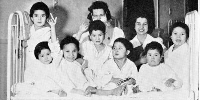 Seven young children sit on a hospital bed. Two women stand behind them.