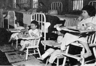 Four children sit in highchairs in front of hospital beds. Older children can be seen sitting on beds in the background.