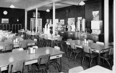 A room filled with dining tables. A cafeteria counter stands agains the right wall. Two women stand behind the counter and two women stand in front.