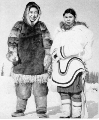 Two people stand in the snow. One person wears a fur coat and the other wears an amautik.