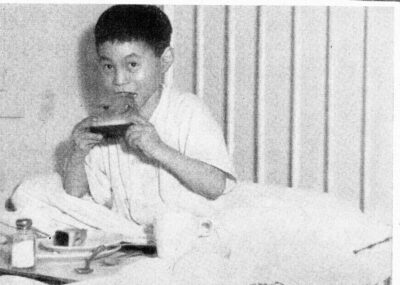 A boy sits on a hospital bed and eats a slice of watermelon. A tray of food sits on the bed in front of him.