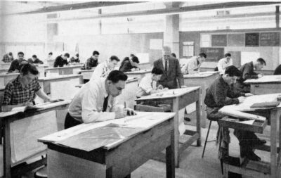 A room filled with men working at drafting tables. A man in a suit stands next to one desk.