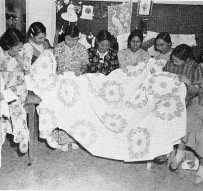 Seven women hold a quilt.