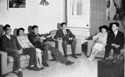 Six smiling people sit on sofas. One man holds a guitar.