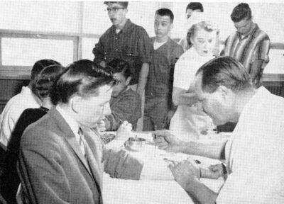 A man holds a tool against another man's forearm. Other people stand around the table, waiting. A nurse looks at the man's forearm.