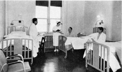 Three male patients sit beside their hospital beds. A nurse stands next to one of the men.