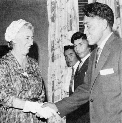 A man in a suit shakes a woman's hand. Two other men stand next to them.