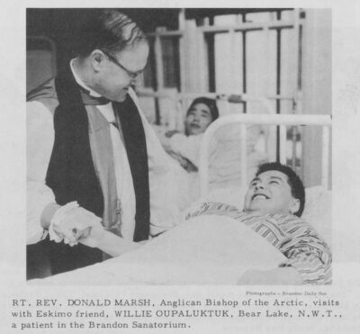 A patient lies in bed and holds the hand of a man who stands next to him. The men smile at each other. Another patient watches from a bed behind them.