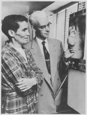 A man in a suit holds a model of a heart as a man in a plaid robe looks at the model. An x-ray image is on the wall in front of them.