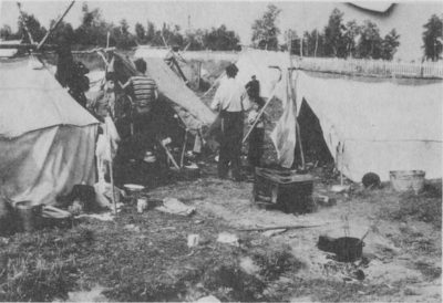 A camp scene with small tents. Some people can be seen amongst the tents.