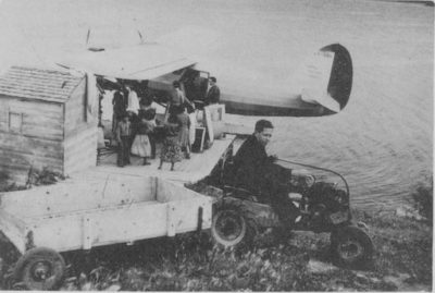 A man on a tractor pulling an open wooden trailer near a docked sea plane. A group of people stand on the dock next to the plane.