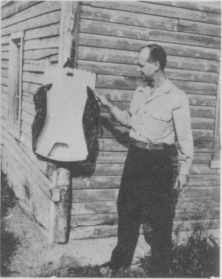 A man stands outside of a log building and places one hand on a moss bag.