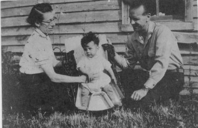 A toddler sits in a chair-like moss bag outdoors, and a man and woman crouch on either side of him.