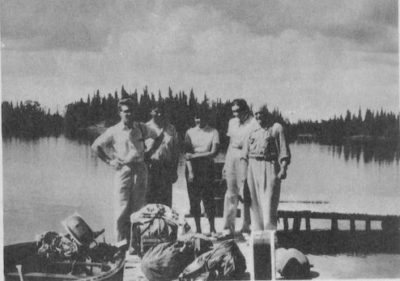 A group of people stand on a dock at a lake. Bags of supplies sit at their feet.