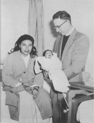 A woman sits on a bed and looks at the camera. A man stands beside her and holds a baby wrapped in a blanket.