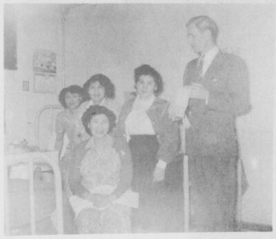 Four women sit on a bed and a man stands next to them.