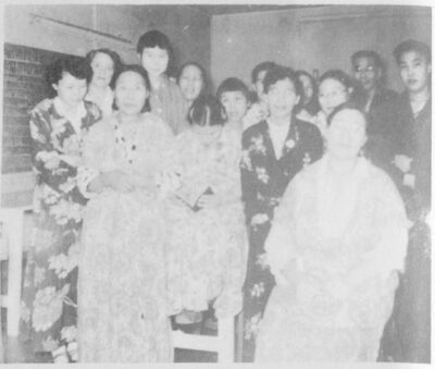 A group of people gathered together. A blackboard can be seen on the wall on the left of the frame. Some people wear floral robes.