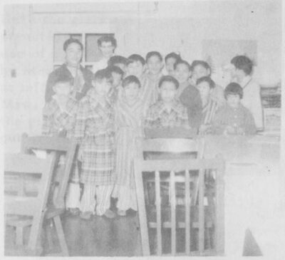 A group of boys gathered at the back of a room. Wooden chairs sit in front of them.