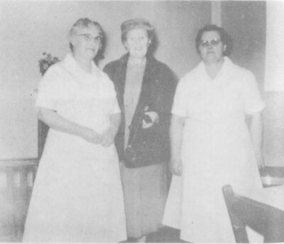 Three women stand together. Two are wearing white dresses.