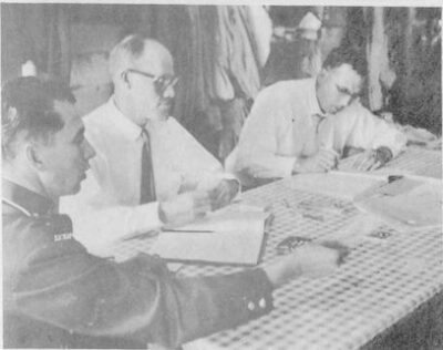 Three men sitting at a table. Two men have open books in front of them, and one man writes in his book.