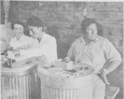 A woman and two men sit behind metal barrels. They are filing and stamping paperwork on top of the barrels.