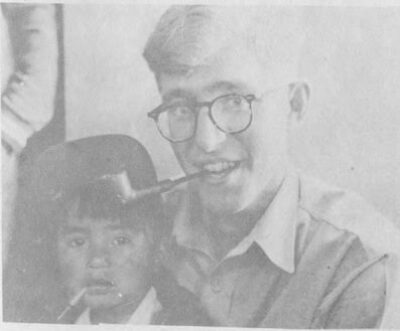 A man with glasses holds a young boy on his lap. They both hold pipes between their teeth.