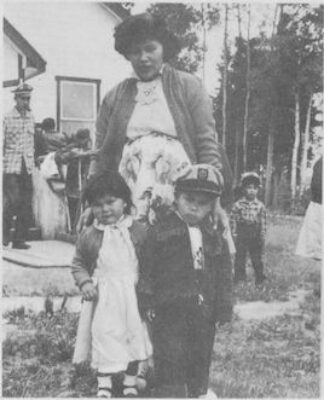 A woman stands with her hands resting on two young children in front of her. Other people can be seen in the background next to a building.