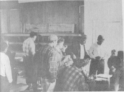 A group of men gathered around seated men writing in books.
