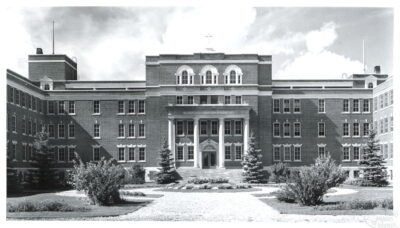 The exterior of a large building. The main entrance is at the centre, and two wings of the building extend towards the viewer on either side.