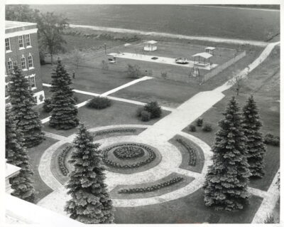 A view from above of the lawn next to a building. There is a circular paved area decorated with flowers, with evergreen trees on the the border. A playground can be seen in the background.