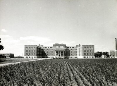 The exterior of a large building, viewed from a distance. A field with rows of plants extends from the bottom of the photo towards the building. Two smaller buildings can be seen on either side.