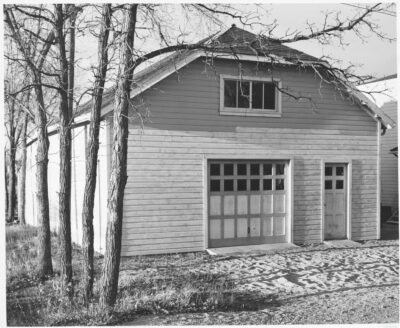 The exterior of a building with a garage door and a regular door. Skinny bare trees stand beside the building.